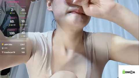 Media: Video of a young East Asian woman with light skin, wearing a beige top, taking a selfie with her left hand covering her mouth, and a digital overlay displaying heart emojis and viewer counts.