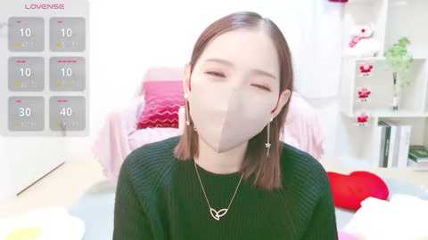 Media: Video of a young woman with straight brown hair, wearing a black sweater, mask, and heart necklace, sitting in a brightly lit bedroom with pink and white decor, including a calendar.