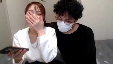 Media: Video: Two East Asian women wearing white masks, seated on a bed. One woman, with long brown hair, covers her face with her hands, looking upset. The other, with curly black hair, is comforting her.