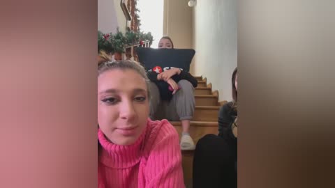 Media: Video of a young woman with light skin and blonde hair, wearing a pink sweater, taking a selfie in a hallway. In the background, another woman sits on stairs, focused on her laptop.