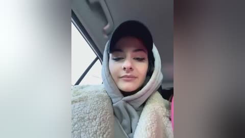 Media: A video of a young woman with light skin, wearing a gray hoodie and dark hair, sitting in a car with a blurry background.