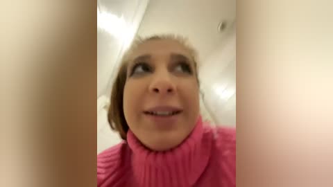 Media: Video of a woman with light skin, brown hair, and pink sweater, looking surprised, with blurry background of white walls and ceiling.