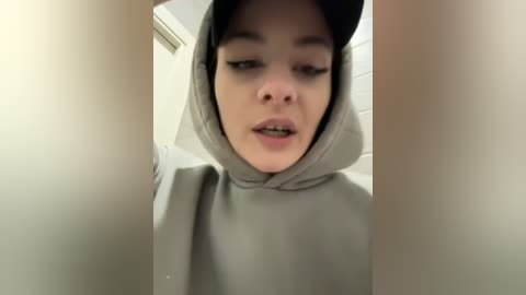 Media: Video of a young person with light skin and straight brown hair, wearing a gray hoodie and black baseball cap, looking through a narrow opening, expression neutral.