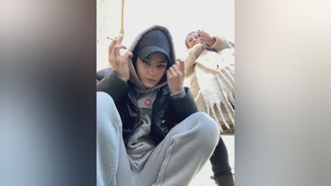 Media: Video of two young Asian women, one squatting with a serious expression, wearing a black hooded jacket, gray sweatpants, and a cap; the other standing, holding a phone, in a beige coat.