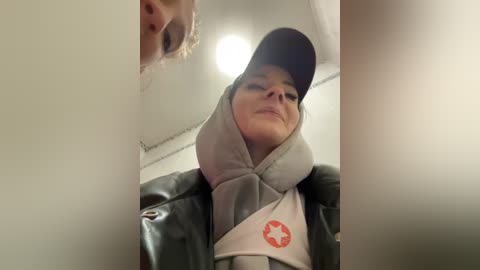 Media: Video of a person wearing a gray hoodie, black baseball cap, and gray jacket, leaning against a white wall with a reflective surface.