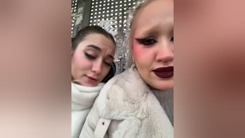 Media: Video of two women, one with a white fur coat, the other with dark lipstick and dramatic eye makeup, standing in front of a silver sequin background.