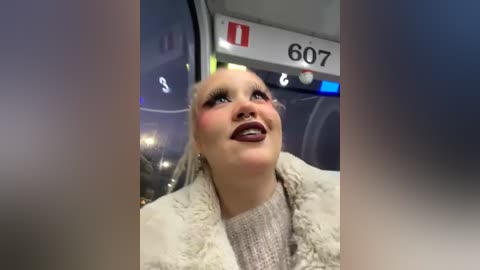 Media: Video of a smiling woman with fair skin, platinum blonde hair, and dark lipstick, wearing a fur coat, inside a train with digital display 607 visible.