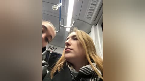 Media: Video of a young blonde woman with long hair, wearing a black coat and a plaid scarf, looking up in a subway train. The background shows the train's interior with fluorescent lights, and the number \"607\" on a sign.