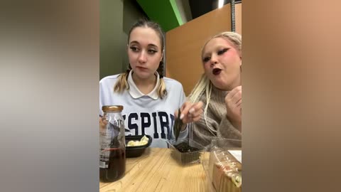 Media: Video of two young women with light skin and long hair, one in a white shirt with \"SPRING\" and the other in a beige top, eating at a wooden table in a dimly lit room with green walls and a metal chair.