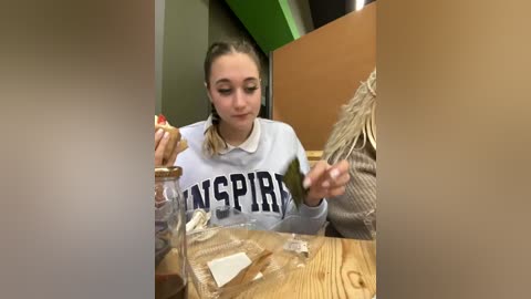 Media: Video of a young woman with light skin and brown hair in a ponytail, wearing a white sweater with \"WINGP\" on it, eating a sandwich. Background shows wooden and green walls.