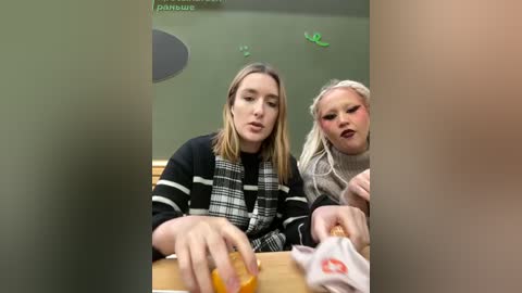 Media: Video of two women with long hair, one in a black plaid shirt, the other in a beige sweater, eating food in a dimly lit, green-wall restaurant.