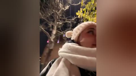 Media: A video of a young woman with fair skin, wearing a white beanie with a bear face, a thick white scarf, and a black jacket, hugging a bare tree in a dark, wintry setting.