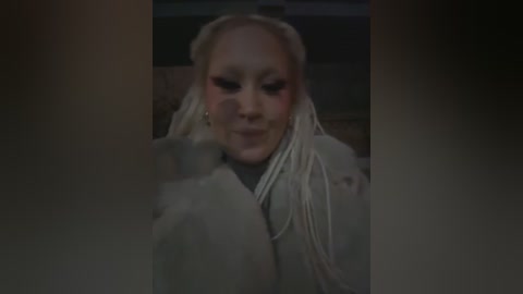 Media: A video of a light-skinned woman with platinum blonde braids, wearing a gray coat, smiling, with a blurred background.