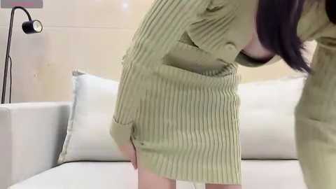 Video of a woman with long dark hair, wearing a green, ribbed, long-sleeved dress, bending over in a modern, minimalist living room with a white couch, a lamp, and beige walls.