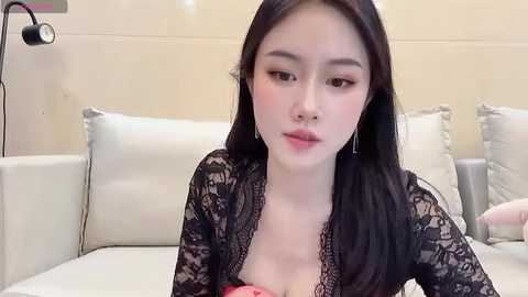 Media: Video of an East Asian woman with fair skin, long black hair, and small breasts, wearing a black lace top and sitting on a white couch.