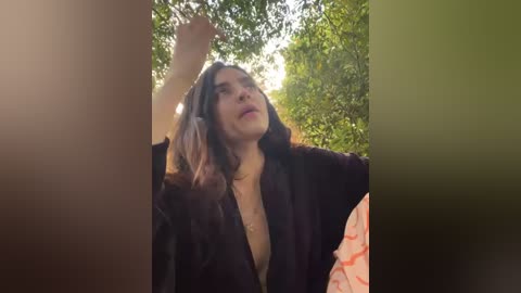 Media: Video of a woman with long, wavy dark hair, wearing a dark, open robe, standing outdoors in a sunlit forest, reaching up.