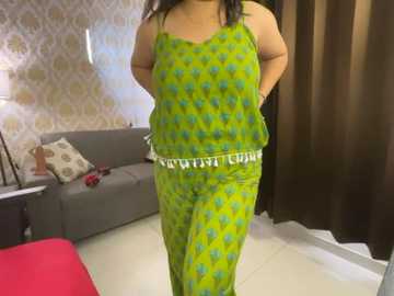 Media: Video of a plus-sized woman in vibrant green pajamas with a \"WOMEN\" waistband, standing in a modern living room with gray couch, patterned wallpaper, and red rug.