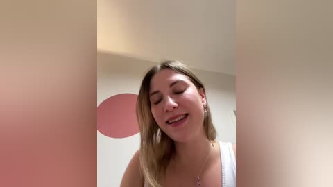 Media: A video of a young Caucasian woman with straight, light brown hair, wearing a white tank top, smiling, standing indoors against a beige wall with a red balloon.