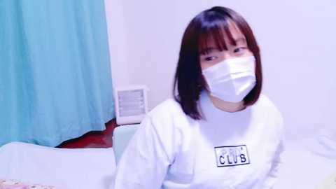 Media: Video of an East Asian woman with shoulder-length black hair, wearing a white mask and \"CLUB\" t-shirt, sitting on a bed with a turquoise curtain in the background.