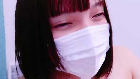 Media: Close-up video of an East Asian woman with straight, shoulder-length black hair and blunt bangs, wearing a white surgical mask, and looking slightly to the side with a neutral expression.