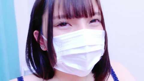 Media: Video of an Asian woman with straight black hair and blunt bangs, wearing a white surgical mask and a blue sleeveless top. Background is a blurred light blue and white.