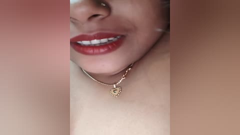 Media: Close-up video of a woman's face, smiling with red lipstick, wearing a gold heart necklace and a thin gold chain. The background is blurred and warm-toned.