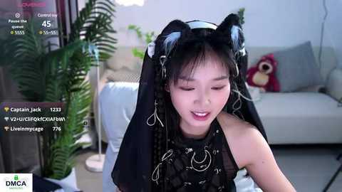 Media: Video of an East Asian woman in a dark, gothic outfit with black cat ears, sitting in a modern living room with plants and a plush toy.