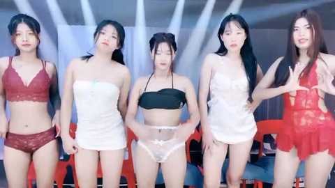 Media: Video of five East Asian women in lingerie, posing in a modern room with white walls and red chairs. They have various hair colors and wear revealing outfits, including lace, satin, and sheer fabrics.