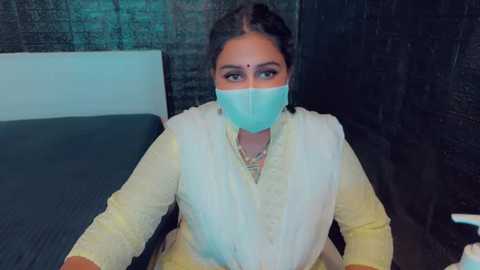 Media: Video of a woman in a yellow saree with a white shawl, wearing a light blue mask, sitting on a bed with a dark headboard and a teal pillow.