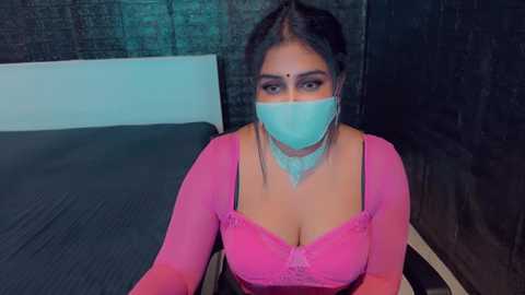 Media: Video of a South Asian woman with medium skin tone, wearing a pink lace bralette, light blue face mask, and dark hair, seated on a bed with dark bedding.