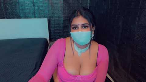 Media: A video of a South Asian woman with medium skin tone, wearing a pink long-sleeve top that reveals cleavage, and a light blue face mask, seated on a bed with dark bedding and a black wall in the background.