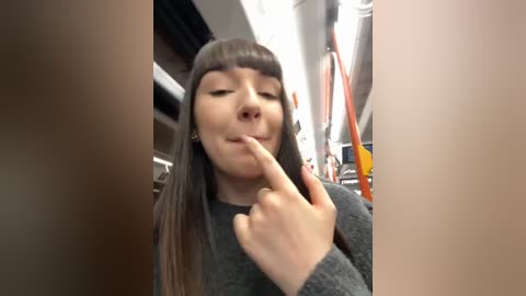 Media: Video of a young woman with long dark hair and bangs, wearing a gray sweater, making a peace sign with her right hand. She is in a subway car with orange poles and overhead lights.