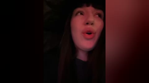 Media: Video of a young woman with long, straight black hair and fair skin, wearing a black hat, looking upward with a surprised expression, in a dimly lit, red-tinted room.