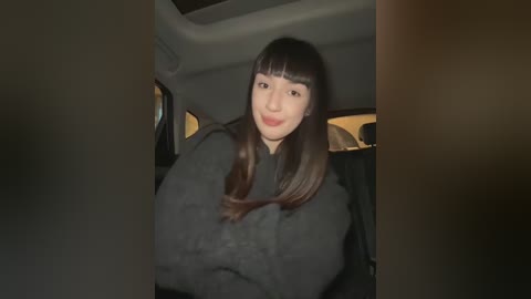 Media: Video of a young Asian woman with long, straight black hair and blunt bangs, wearing a black sweater, smiling inside a dimly lit car.