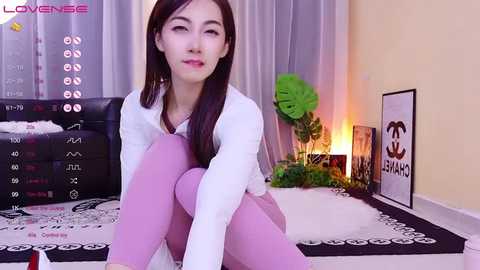 Media: Video of an Asian woman with straight black hair, fair skin, and pink lipstick, wearing a white blouse and lavender pants, sitting on a bed with black and white bedding, surrounded by green plants and a framed Chanel logo.