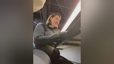 Media: Video of a woman with shoulder-length brown hair, wearing a grey zip-up jacket and black leggings, working on a large white structure in a dimly lit, industrial space.