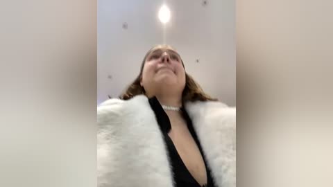 Media: Video of a woman with fair skin, wearing a black dress and a white fur coat, standing in a modern room with a spotlight shining on her face.