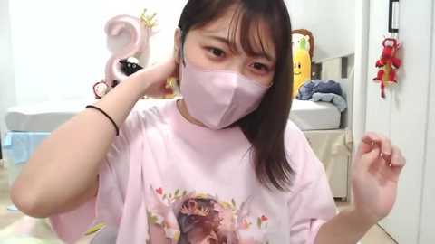 A video of an Asian woman with light skin, dark hair, and a pink face mask, wearing a graphic t-shirt, in a brightly lit room with pastel decor and plush toys.