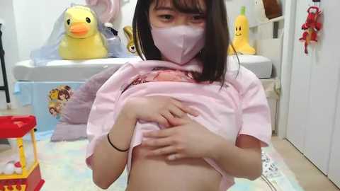 Video of an East Asian woman with straight black hair, wearing a pink mask, lifting her pink T-shirt to reveal her small breasts, in a playroom with toys and plushies.