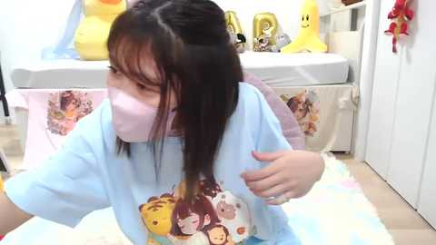 Media: Video of a young Asian woman with long black hair, wearing a light blue T-shirt featuring anime characters, pink face mask, and white gloves, seated in a brightly lit, plush bedroom with toys and a bed in the background.