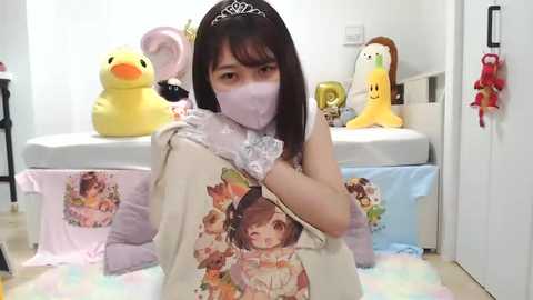 Video of an Asian woman with long black hair, wearing a white face mask, a pastel floral shirt, and a yellow headband, surrounded by plush toys in a bright, clean room.