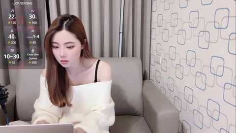 Media: Video of a young East Asian woman with long brown hair, wearing a white off-shoulder sweater and black bra, typing on a laptop in a modern living room with patterned wallpaper and gray curtains.