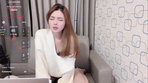 Media: Video of an Asian woman with straight, light brown hair, wearing a white top, sitting on a gray sofa, eyes closed, in a room with geometric-patterned wallpaper and a digital clock displaying 6:00 PM.