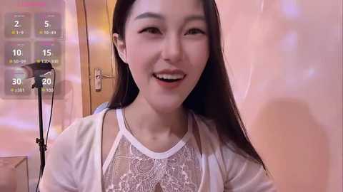 Media: Video of a smiling East Asian woman with long black hair, wearing a white lace top, in a cozy room with a calendar on the wall.