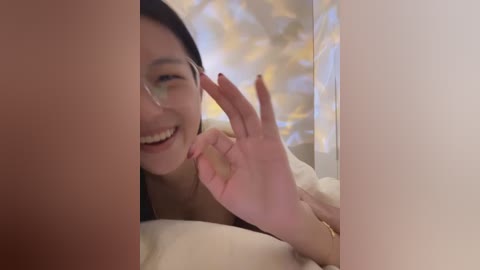 Media: Video of an Asian woman with long black hair, wearing glasses, smiling, making an OK sign with her right hand, lying in bed with white sheets, blurred background.