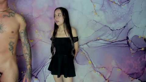 Media: Video of a tattooed man and a young woman with long black hair, wearing a black off-shoulder dress. They stand in front of a marbled purple and white backdrop.