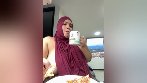 A video of a woman in a burgundy hijab and sleeveless top, holding a white mug, eating pizza in a modern, bright kitchen with a view of a cityscape.