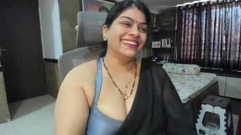 Media: A video of a South Asian woman with medium skin tone, black hair, and a warm smile. She wears a blue tank top and a black shawl, sitting on a grey chair in a modern bedroom with a bed, curtains, and shelves in the background.