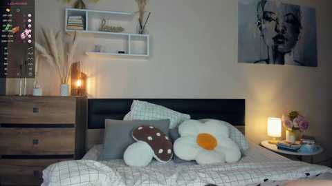 Media: Video of a minimalist, cozy bedroom with a wooden dresser, white shelves, a large floral pillow, a lit lamp, and a monochromatic portrait on the wall.