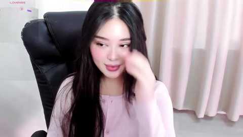 Media: A video of an East Asian woman with long black hair, fair skin, and pink lipstick, wearing a light pink cardigan, seated in a black leather chair. The background features white walls and a pink curtain.